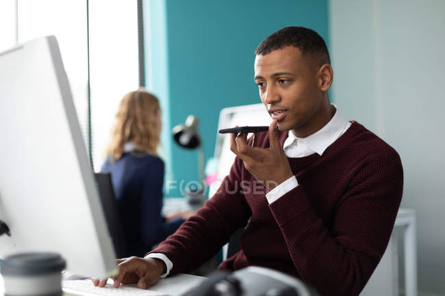 Вид спереди на молодого афроамериканца, сидящего за столом, разговаривающего на смартфоне в современном офисе творческого бизнеса, со своей коллегой-женщиной, работающей на заднем плане — стоковое фото