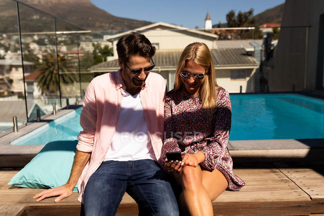 Vista frontal de um jovem casal caucasiano feliz relaxando de férias sentado à beira de uma piscina, usando um smartphone — Fotografia de Stock