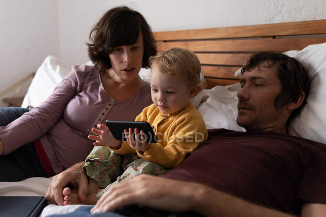 Вид сбоку: молодой кавказский отец и мать лежат на кровати, в то время как их ребенок держит смартфон — стоковое фото