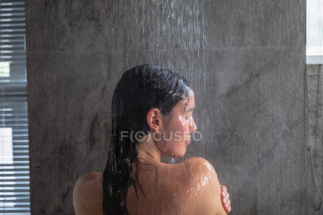Закрити заднього виду молодої кавказької жінкою брюнетки стоячи в душі мити волосся, з її голова перетворилася на бік у сучасній ванній кімнаті — стокове фото