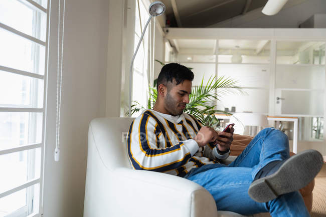 Vista laterale da vicino di un giovane uomo di razza mista seduto su una poltrona e che utilizza uno smartphone in un ufficio creativo — Foto stock