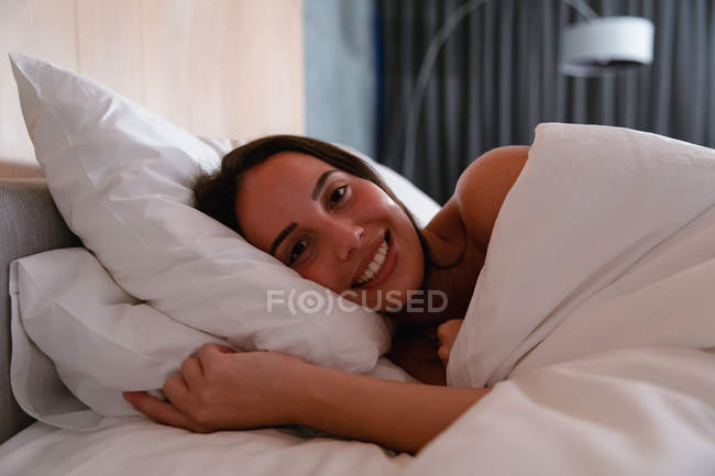 Retrato de cerca de una joven morena caucásica acostada de costado en la cama sonriendo a la cámara - foto de stock