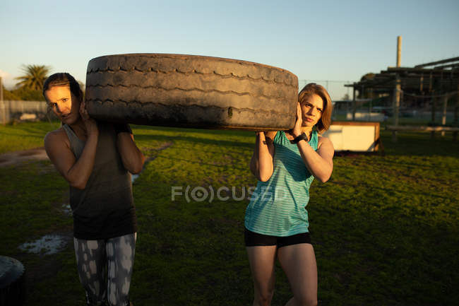 Vista frontal de duas jovens mulheres caucasianas carregando um pneu em um ginásio ao ar livre durante uma sessão de treinamento de bootcamp — Fotografia de Stock
