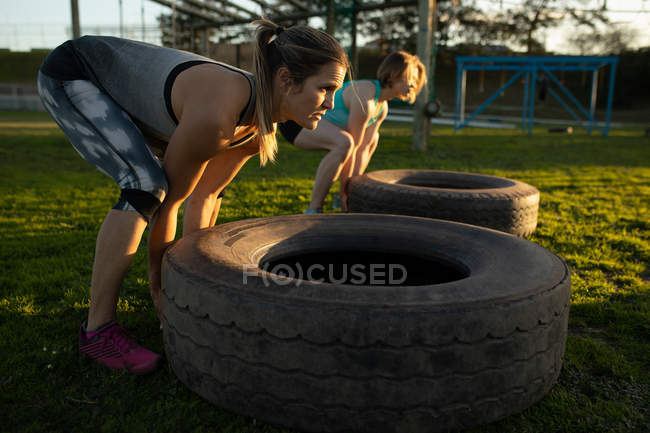 Vista lateral de dos jóvenes mujeres caucásicas volteando neumáticos en un gimnasio al aire libre durante una sesión de entrenamiento de bootcamp - foto de stock