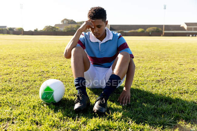 Vista frontal de cerca de una joven adulta de raza mixta jugadora de rugby sentada en un campo de rugby en pensamiento, con la pelota a su lado - foto de stock