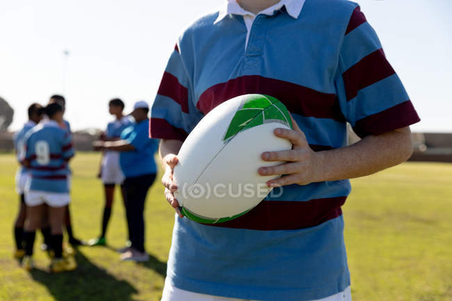 Vue de face section médiane d'une jeune joueuse de rugby blanche adulte debout sur un terrain de rugby tenant une balle de rugby, avec ses coéquipières parlant ensemble en arrière-plan — Photo de stock
