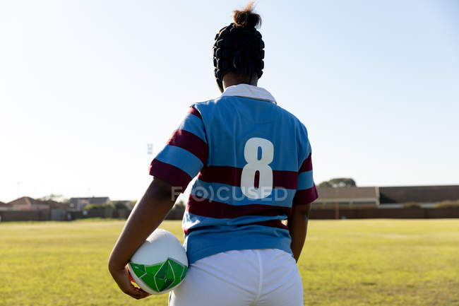 Вид сзади на молодую взрослую регбистку смешанной расы в костюме охранника, стоящего на поле для регби с мячом для регби — стоковое фото