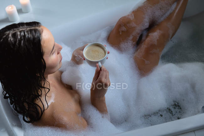 Vista elevada de una joven morena caucásica sentada en un baño de espuma con los pies en alto sosteniendo una taza de café - foto de stock