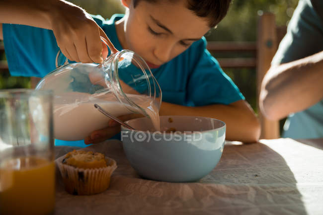 Vista frontal close-up de um menino pré-adolescente caucasiano sentado em uma mesa desfrutando de um café da manhã em família em um jardim, derramando leite em uma tigela — Fotografia de Stock