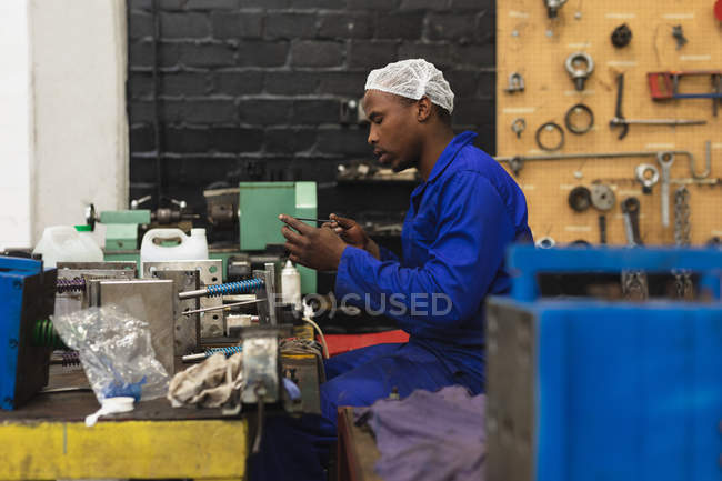 Vista lateral de cerca de un joven trabajador de la fábrica afroamericano sentado e inspeccionando equipos en el taller de máquinas en una planta de procesamiento, con equipos y herramientas en el fondo - foto de stock