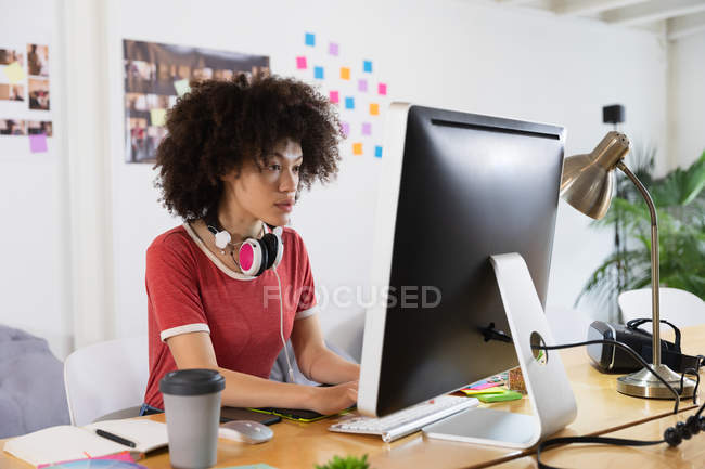 Side view of a young mixed race woman sitting at a desk typing and looking at a computer screen at a creative office — Stock Photo