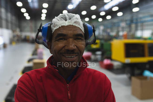 Portrait en gros plan d'un ouvrier afro-américain d'âge moyen habillé de vêtements de travail avec des protecteurs d'oreilles sur la tête, debout dans un entrepôt dans une usine de transformation souriant à la caméra avec les bras croisés — Photo de stock