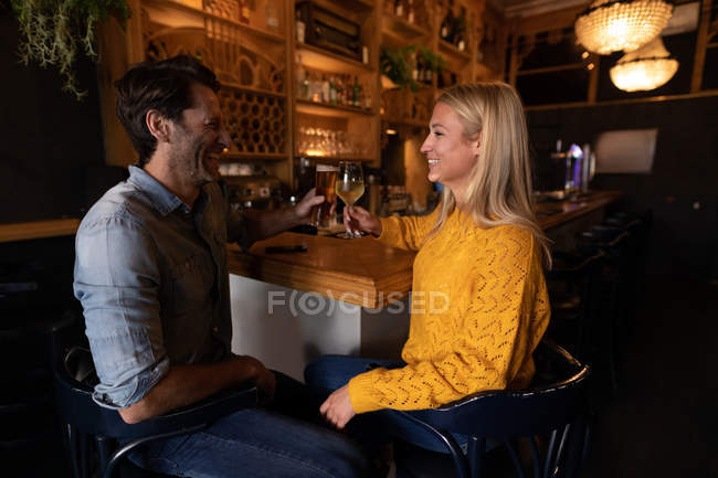 Вид спереди на счастливую молодую кавказскую пару, отдыхающую вместе в баре, пьющую пиво, вино и улыбающуюся — стоковое фото