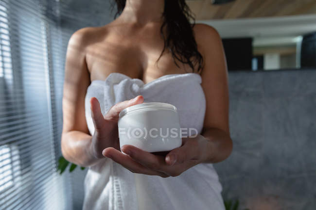 Вид спереду середня секція жінки в рушнику для ванни, що тримає банку красивого крему в сучасній ванній кімнаті — стокове фото