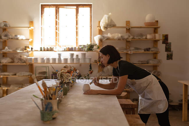 Vista laterale di una giovane ceramista caucasica appoggiata su un tavolo da lavoro e che invetriava una pentola in uno studio di ceramica — Foto stock