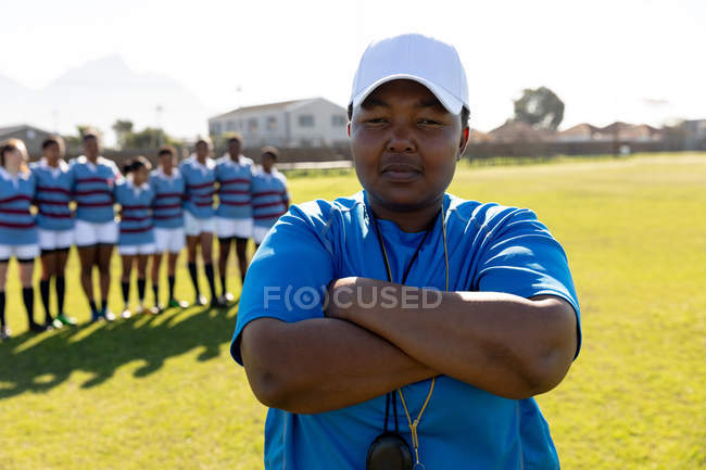 Portrait close up of a middle aged mixed race female rugby coach standing on a rugby pitch with her arms crossed looking to camera, with her team standing in a row in the background — Stock Photo