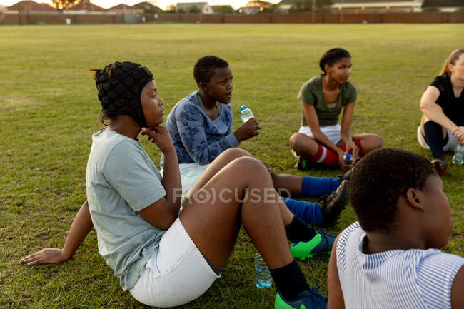 Вид сбоку команды молодых взрослых многоэтнических регбисток, сидящих на регбийном поле во время тренировки — стоковое фото