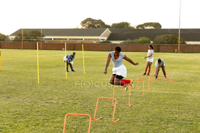 Вид спереди на молодого взрослого игрока смешанной расы, прыгающего с препятствиями на регбийном поле во время тренировки, с товарищами по команде на заднем плане — стоковое фото