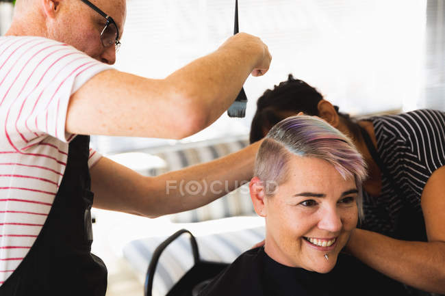 Front view close up of a middle aged Caucasian male hairdresser, a middle aged mixed race female hairdresser and a young Caucasian woman having her hair coloured in a hair salon — Stock Photo