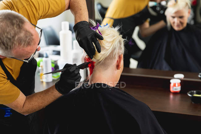 Visão traseira close-up de um cabeleireiro masculino caucasiano de meia idade e uma jovem mulher caucasiana com seu cabelo colorido vermelho brilhante em um salão de cabeleireiro, refletido em um espelho — Fotografia de Stock