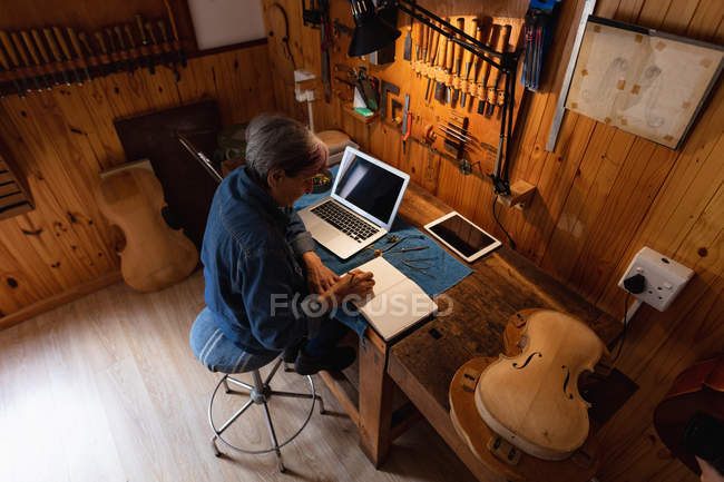 Vista de alto ángulo de una luthier mujer blanca mayor haciendo notas en su taller, con una computadora portátil y una tableta delante de ella y herramientas colgando en la pared en el fondo - foto de stock