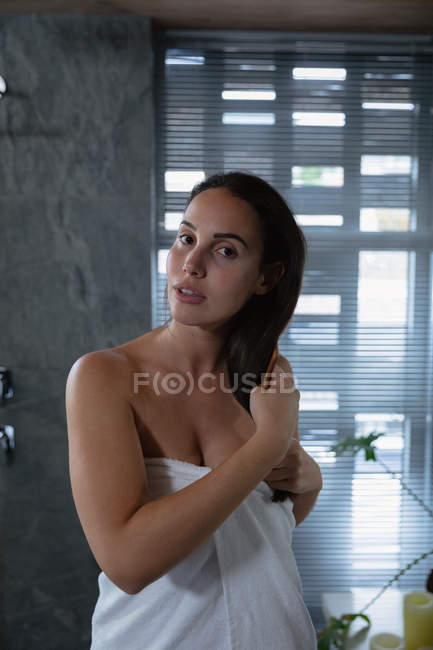 Portrait close up of a young Caucasian brunette woman wearing a bath towel brushing her hair in a modern bathroom — Stock Photo