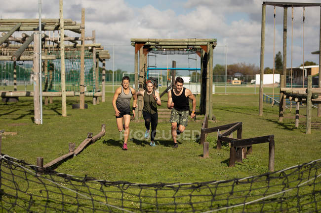Вид спереди на двух молодых кавказских женщин и молодого кавказца, бегущего между препятствиями в спортзале на открытом воздухе во время тренировочного лагеря — стоковое фото