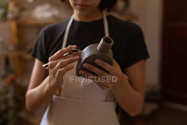 Vista frontal sección central de una joven alfarera caucásica sosteniendo un matraz de arcilla y una herramienta de modelado en un estudio de cerámica - foto de stock