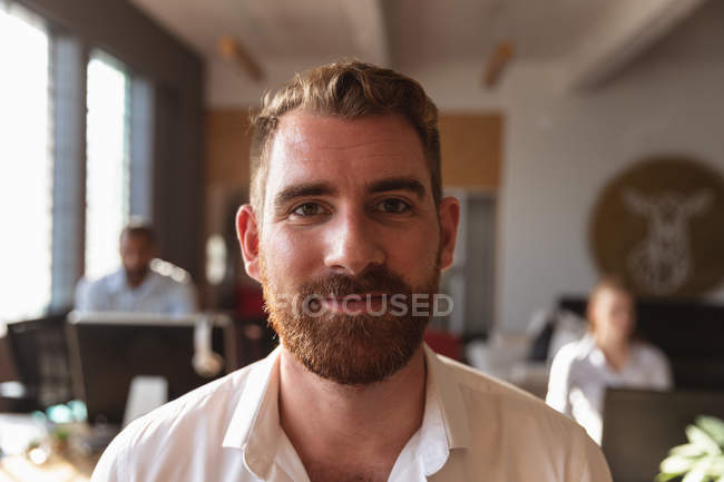 Portrait close up of a young Caucasian man with short hair and a beard looking to camera smiling in a creative office, with colleagues working at desks in the background — Stock Photo