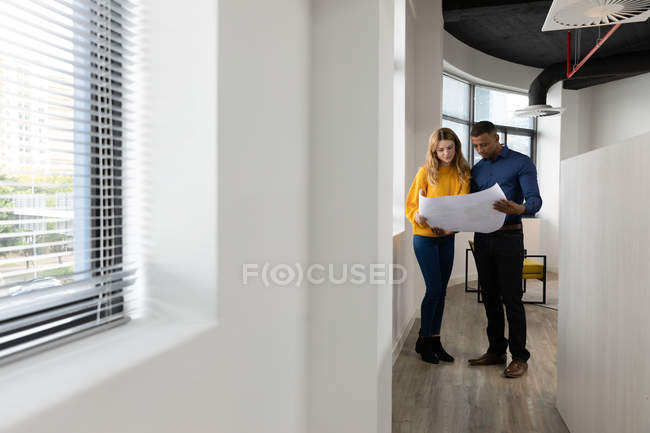 Vista frontale di un giovane uomo afroamericano e una giovane donna caucasica che guardano insieme i piani nel corridoio di un moderno business creativo — Foto stock