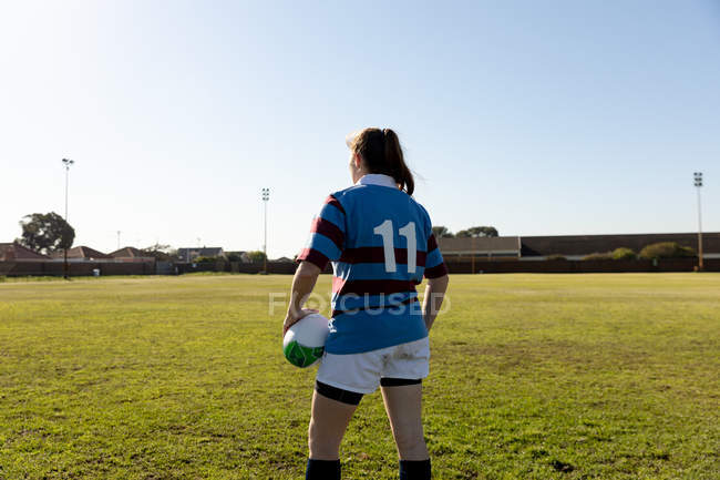 Вид сзади на молодую взрослую белую регбистку, стоящую на поле для регби, держа регбийный мяч — стоковое фото