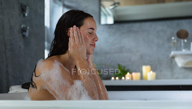 Side view close up of a young Caucasian brunette woman sitting in a bath with lit candles on the side, washing her face with her eyes closed — Stock Photo