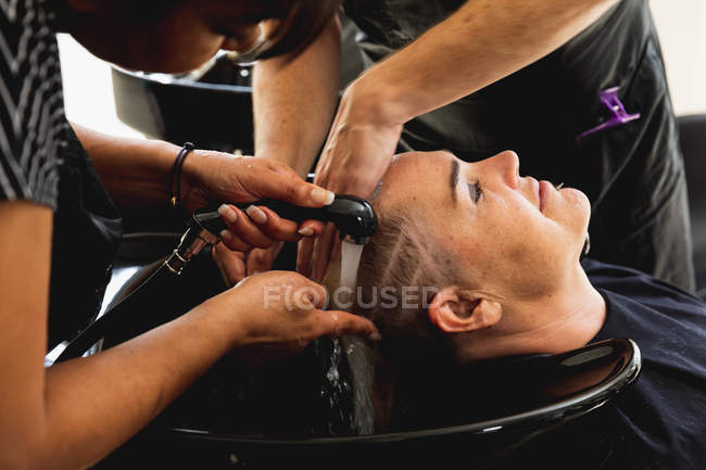 Vue de côté gros plan d'un coiffeur masculin caucasien d'âge moyen, d'un coiffeur féminin métis d'âge moyen et d'une jeune femme caucasienne se faisant laver les cheveux dans un salon de coiffure — Photo de stock