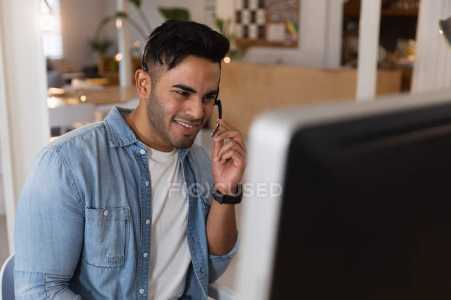 Передній вид крупним планом молодий змішаний чоловік, сидячи за столом в гарнітурі і дивлячись на екран комп'ютера в творчому офісі — стокове фото