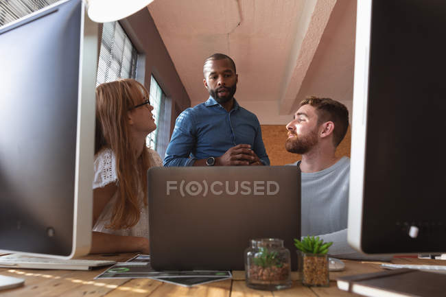 Vue de face gros plan d'un jeune Afro-Américain tenant une tasse de café parlant avec une jeune femme caucasienne et des collègues masculins assis à un bureau à l'aide d'un ordinateur portable dans un bureau créatif — Photo de stock