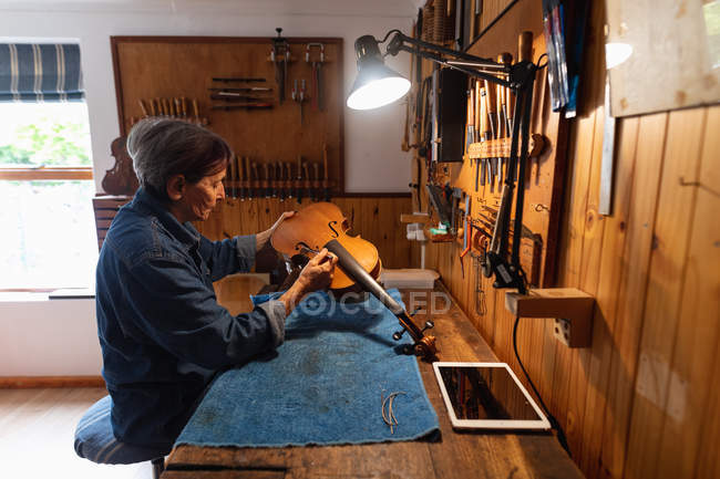 Side view of a senior Caucasian female luthier working on the body of a violin in her workshop, with a tablet computer in front of her and tools hanging up on the wall in the background — Stock Photo