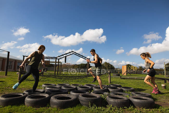 Vista lateral de dos jóvenes mujeres caucásicas y un joven hombre caucásico pisando neumáticos en un gimnasio al aire libre durante una sesión de entrenamiento de bootcamp - foto de stock