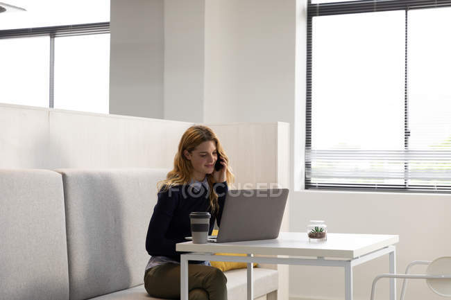 Vorderansicht einer jungen kaukasischen Frau, die mit einem Laptop auf einer Bank sitzt und an einem Tisch mit einer Tasse Kaffee im Essbereich eines kreativen Unternehmens auf einem Smartphone spricht — Stockfoto