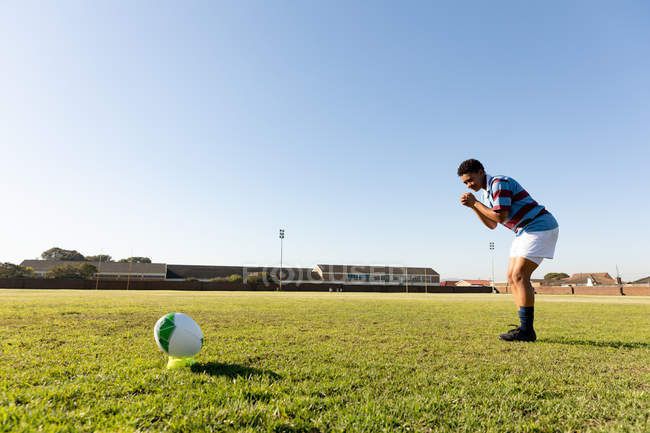 Vista lateral de una joven jugadora de rugby de raza mixta adulta parada en un campo de rugby preparándose para correr hasta la pelota y hacer una patada en el lugar - foto de stock