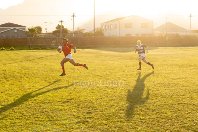 Vista lateral de una joven jugadora de rugby caucásica adulta corriendo tras una joven jugadora de rugby mixta adulta sosteniendo la pelota durante un partido de rugby - foto de stock