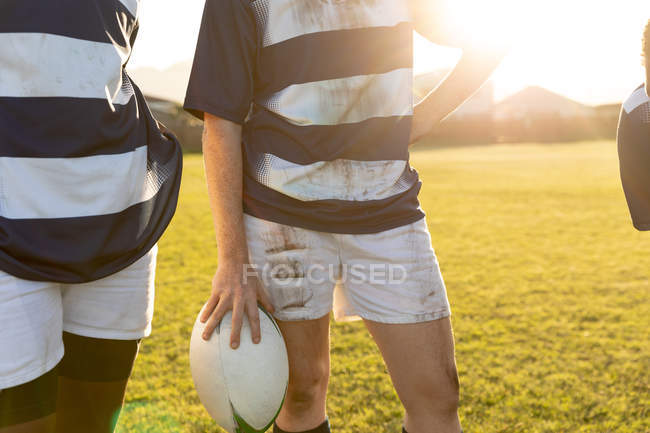 Vista frontal de la sección central de la jugadora femenina de rugby sosteniendo la pelota relajándose después de un partido de rugby, con su compañero de equipo de pie junto a ella - foto de stock
