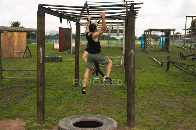 Rückansicht eines jungen kaukasischen Mannes, der während eines Bootcamp-Trainings an einer Affenstange in einem Outdoor-Fitnessstudio hängt, mit anderen Teilnehmern im Hintergrund — Stockfoto