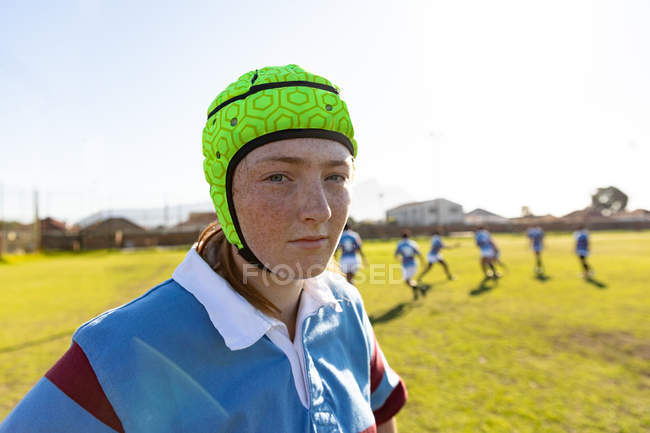 Portrait close up of a young adult Caucasian female rugby player wearing a headguard standing on a rugby pitch, with her teammates in the background — Stock Photo