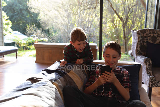Вид спереди двух мальчиков дошкольного возраста, использующих смартфон и слушающих музыку с включенными наушниками в гостиной дома — стоковое фото