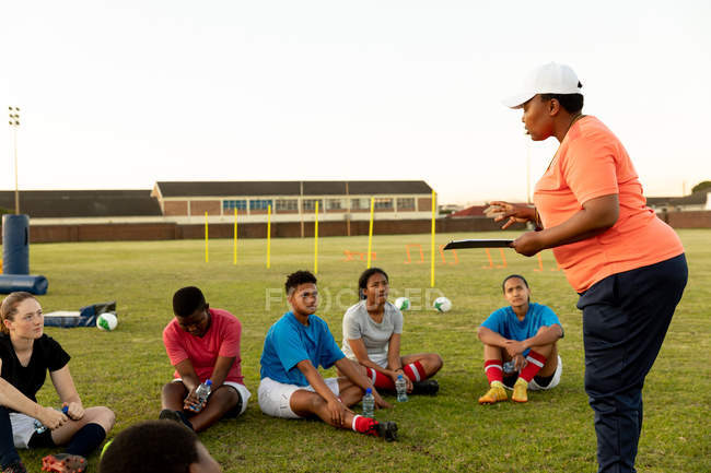Side view of a middle aged mixed race female rugby coach holding a clipboard standing and talking to a team of a young adult multi-ethnic female rugby players sitting on a rugby field during a training session — Stock Photo