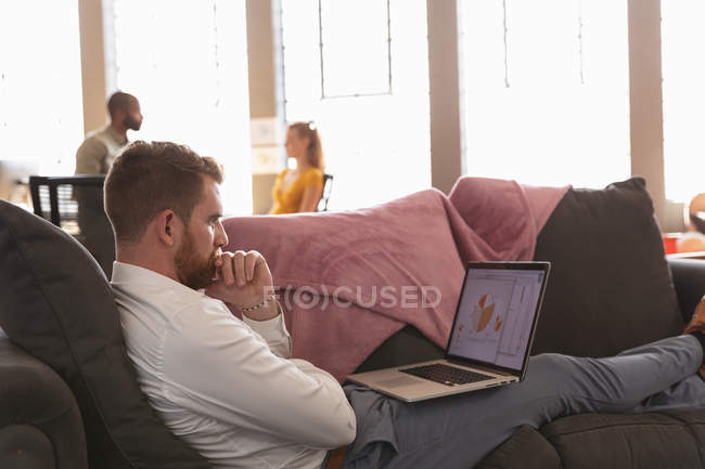 Vista lateral de cerca de un joven hombre caucásico sentado en un sofá con los pies en alto usando una computadora portátil en la sala de estar de una oficina creativa, con un colega trabajando en el fondo - foto de stock