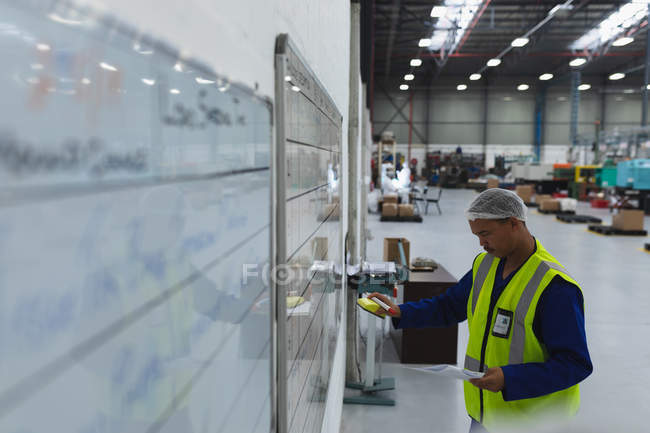 Seitenansicht eines männlichen Fabrikarbeiters mittleren Alters mit Dokumenten, die Informationen auf einem Whiteboard in einer Lagerhalle eines Verarbeitungsbetriebs aktualisieren — Stockfoto