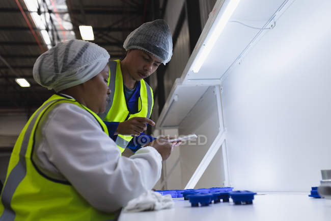 Vue de côté gros plan d'un jeune ouvrier d'usine mixte utilisant un ordinateur tablette parlant avec une jeune ouvrière afro-américaine assise et inspectant des pièces en plastique sous une lumière vive dans un entrepôt d'une usine — Photo de stock
