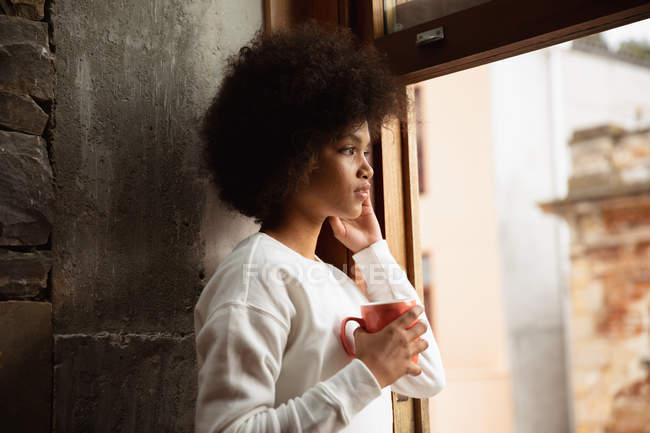 Vista lateral close-up de uma jovem mulher de raça mista em pé por uma janela e olhando para fora segurando uma xícara de café — Fotografia de Stock
