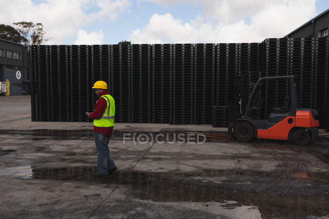 Side view of a middle aged Caucasian male factory worker using a smartphone standing beside a forklift truck and stacks of pallets outside a warehouse at a factory — Stock Photo